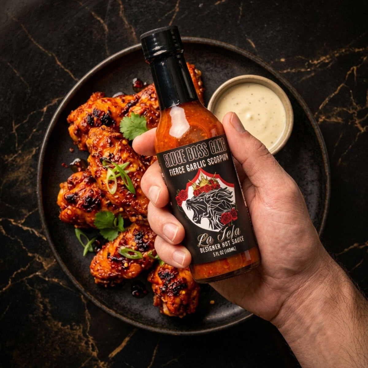 Hand holding La Jefa garlic scorpion hot sauce beside a plate of charred chicken wings